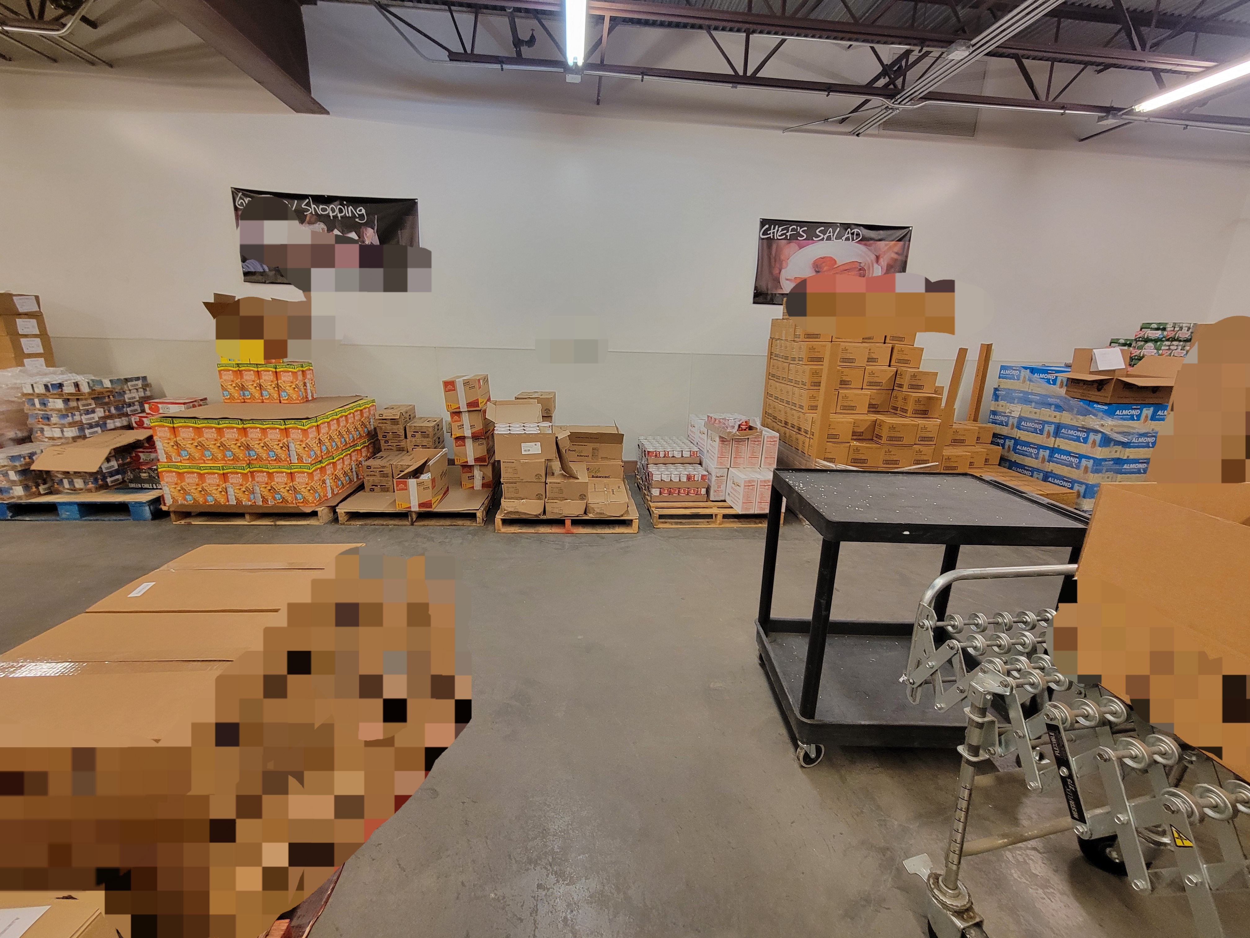 Pallet of boxes to left, and a shelf cart next to a box filled conveyor belt with pallets of food behind it.